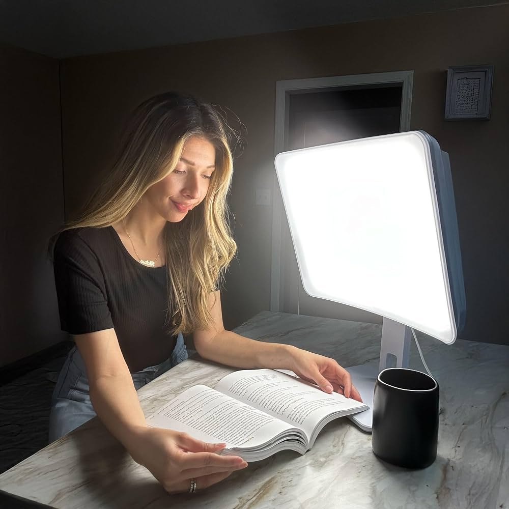 A woman reading at a marble counter beside a Carex Day-Light Sky bright light therapy lamp