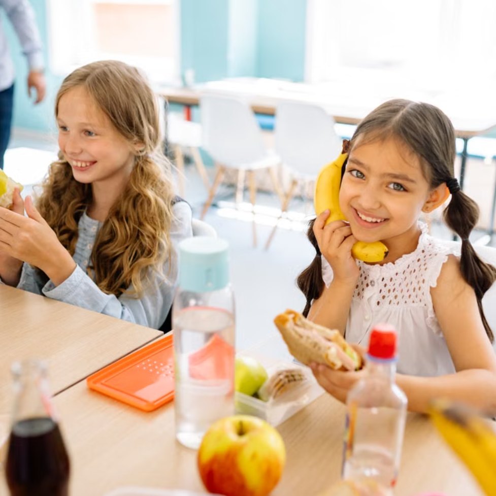 Two schoolchildren at a lunch table with fresh fruit, one holding a pear and one a banana
