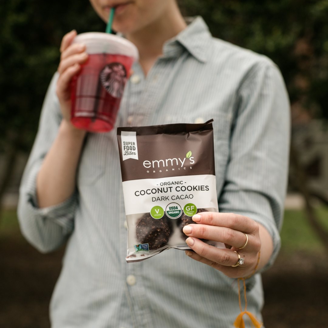 A woman sipping an iced Starbucks drink while holding out a bag of Emmy&rsquo;s Organics Dark Cacao Coconut Cookies