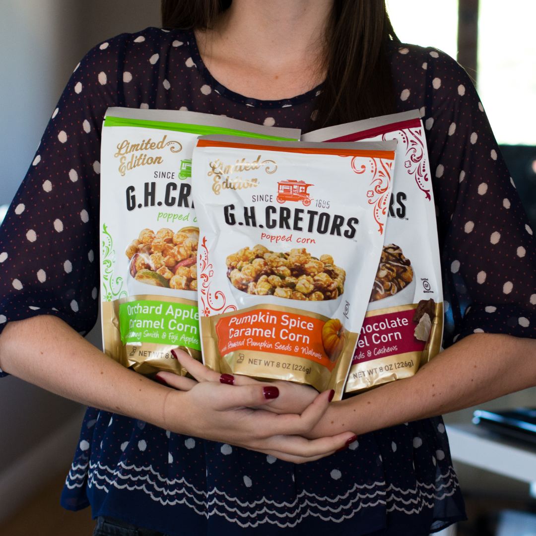 A woman holding three bags of G.H. Cretors limited-edition popped corn: Orchard Apple Caramel Corn, Pumpkin Spice Caramel Corn and Chocolate Caramel Corn