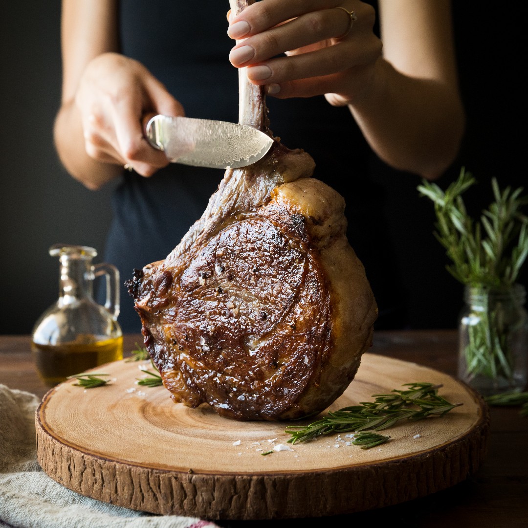A bone-in tomahawk ribeye being carved on a wooden board with rosemary and olive oil