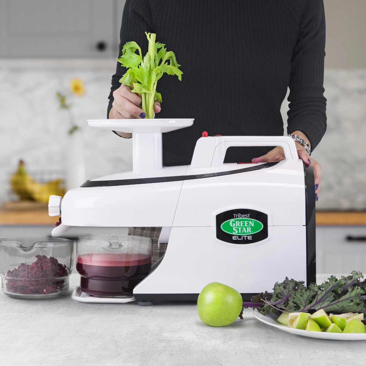 A Tribest Green Star Elite juicer on a kitchen counter with celery, apple and kale alongside fresh juice